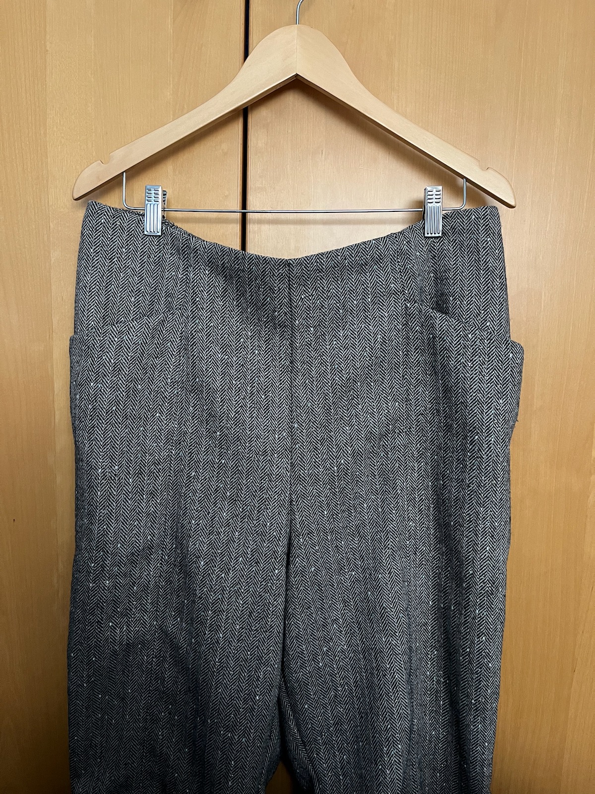 A pair of grey woollen trousers with centre front leg seams and angled pockets on a hanger.