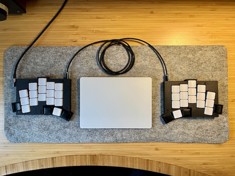 Wired version of TOTEM split keyboard with black case, sitting on grey felt desk mat with Apple Trackpad between the two halves.
