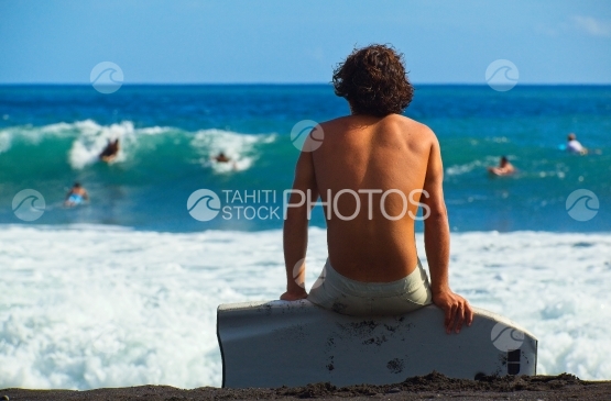 Tahiti, young man sat on his surf board on the beach  watching surfers in the waves