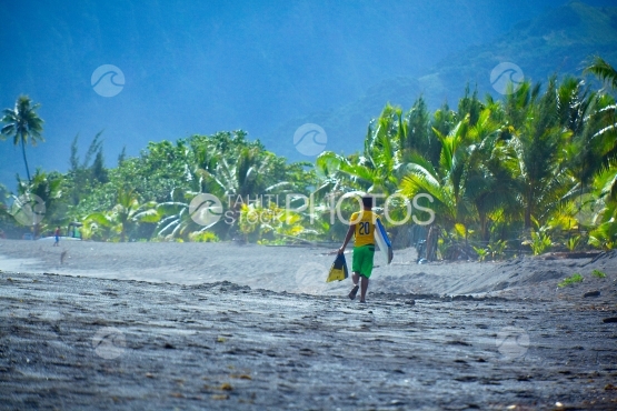 Tahiti, young surfer boy walking on the black sand beach, with his fins and body board