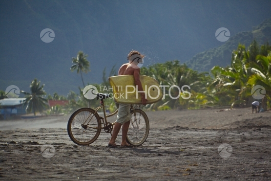 Tahiti, young surfer boy walking on the black sand beach, with his bicycle